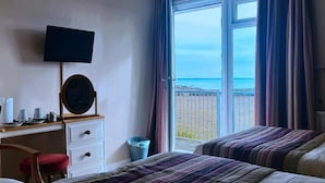 Een hotelkamer met een bed, een bureau met een televisie, een spiegel en uitzicht op de zee door een groot raam.