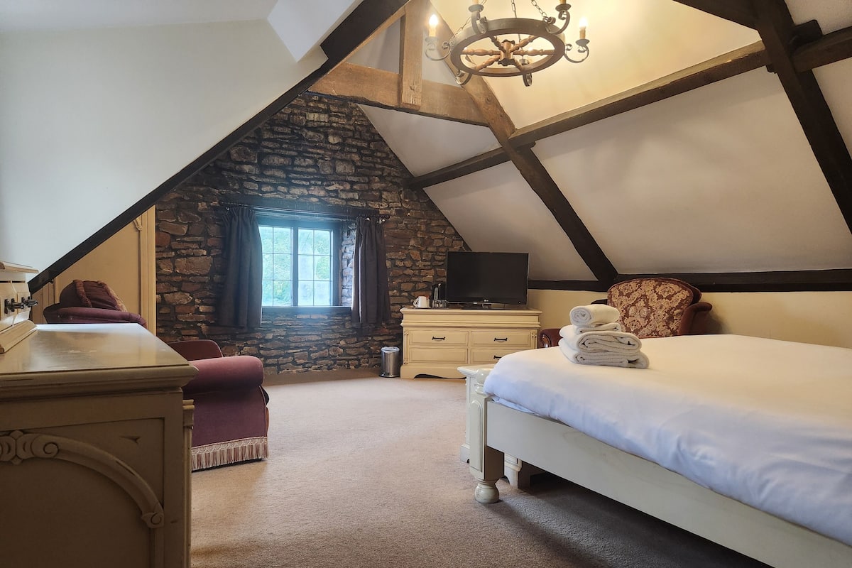 A cozy attic bedroom with a stone wall, a large window, a bed with rolled towels, a TV, and a desk.