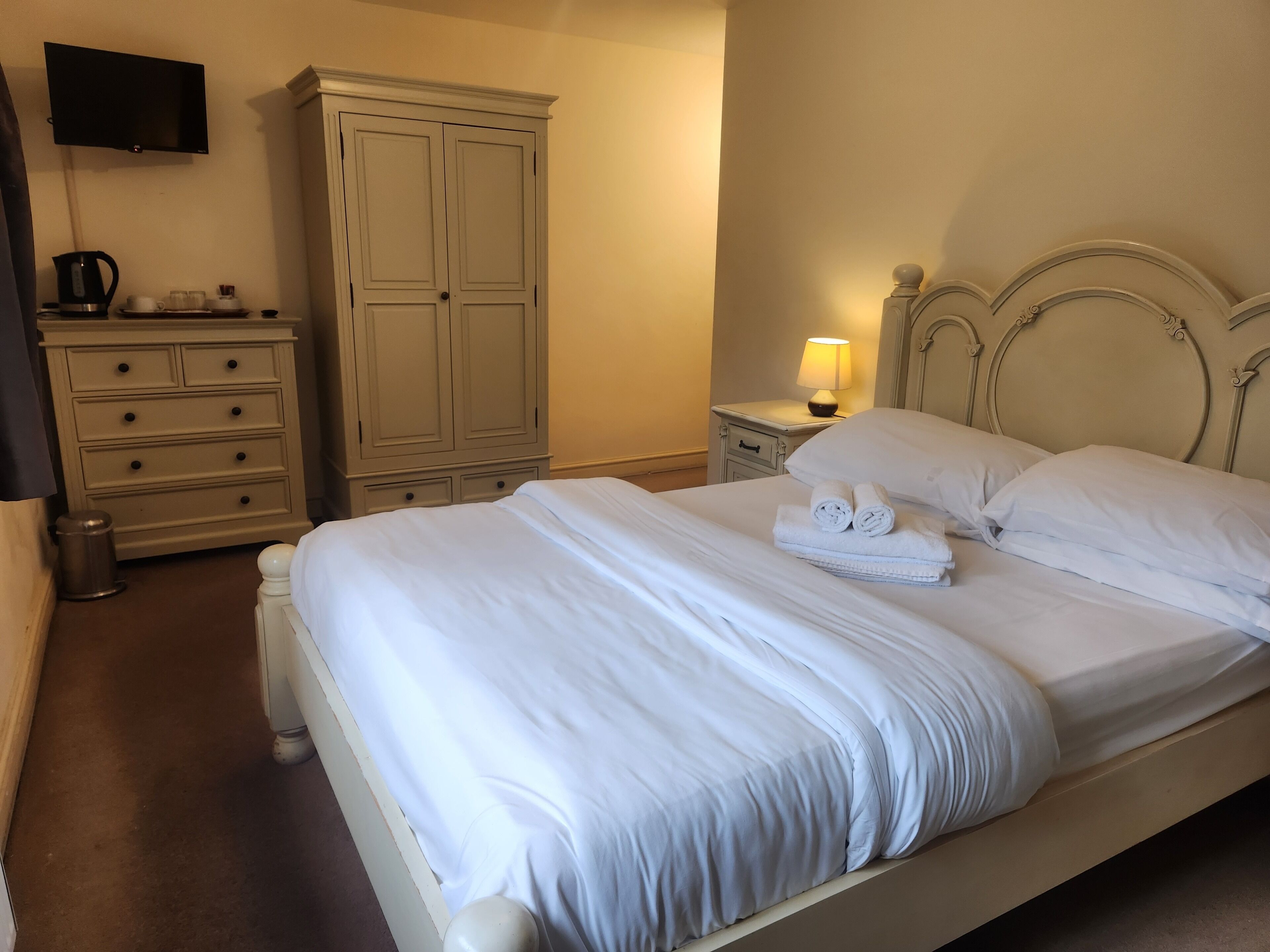 Una cama bien hecha con sábanas blancas y toallas enrolladas, una lámpara de noche, un armario y un televisor montado en la pared.
