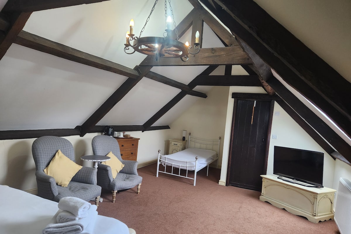 A cozy attic bedroom with a bed, two armchairs, a small table, a dresser, a bedside table, a TV, and a chandelier.