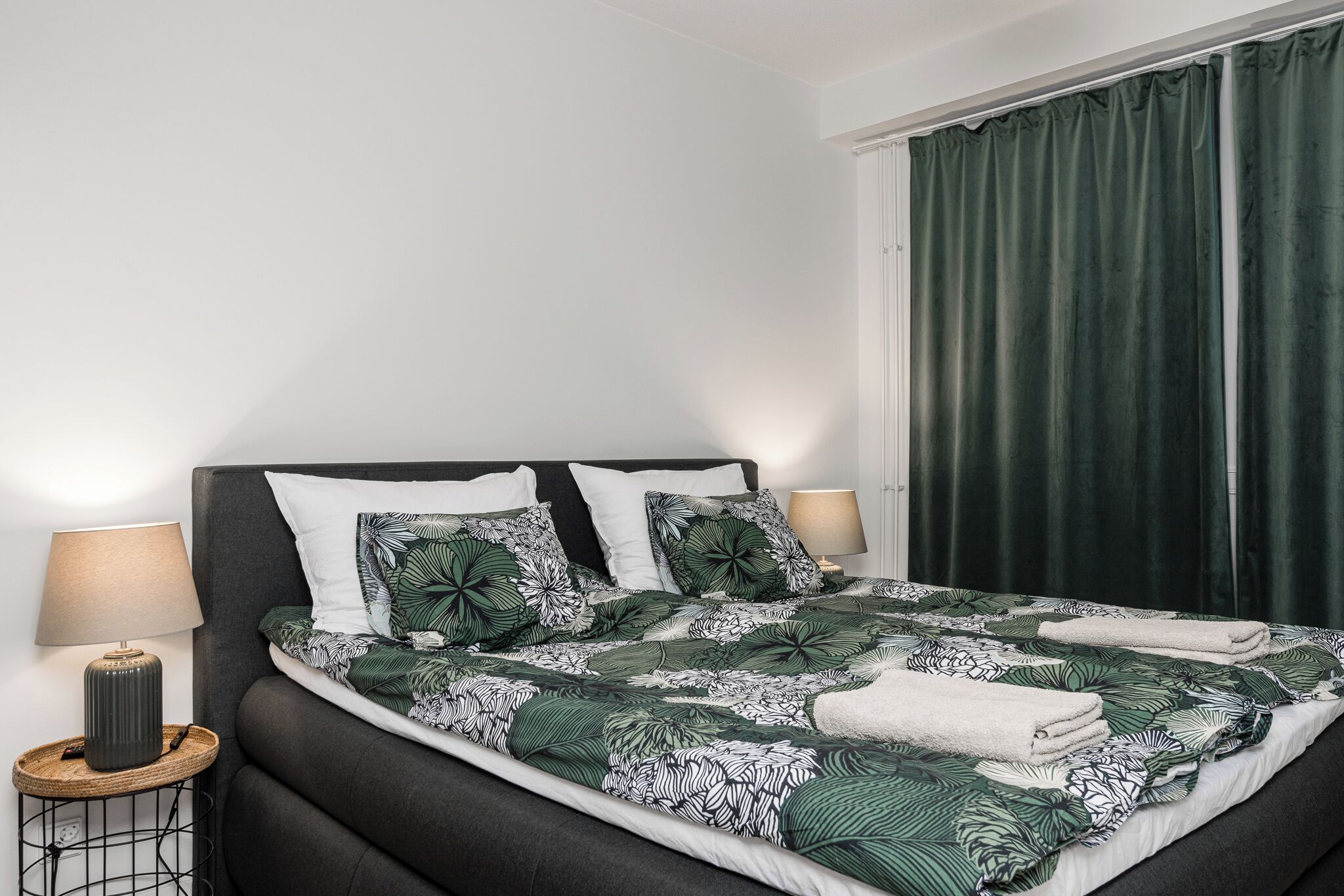 Una cama bien hecha con colcha de estampado tropical, dos lámparas de noche y una cortina verde oscuro.
