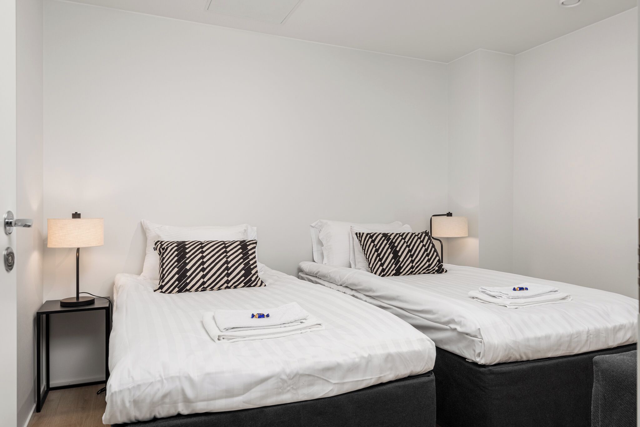 Una habitación de hotel con dos camas individuales, cada una con un cojín a rayas blanco y negro y una colcha blanca. Hay una mesita de noche con una lámpara a cada lado de las camas.