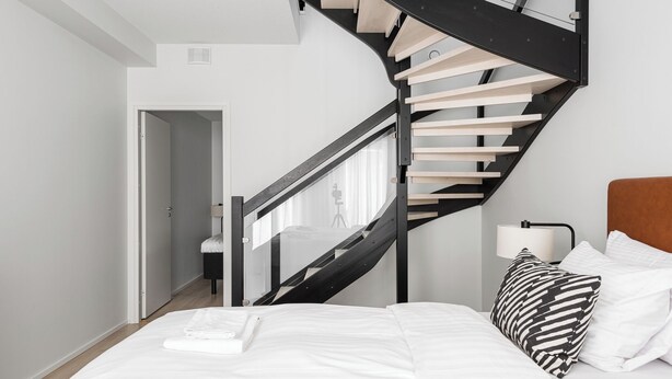 A bedroom with a modern design, featuring a curved staircase, a bed with white bedding, and a nightstand with a lamp. - Itäpuisto Loft apartments by Borg (Pori)