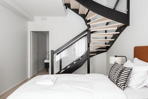 Quarto com design moderno, com escadaria sinuosa, cama com roupa de cama branca e mesa de cabeceira com abajur.