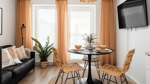 A modern living room with a black leather sofa, a round dining table with wicker chairs, a potted plant, and a large TV mounted on the wall. - Itäpuisto Loft apartments by Borg (Pori)