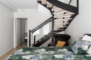 Un dormitorio con una escalera curva, una cama con colcha floral, un sillón de cuero negro y una mesita de noche.