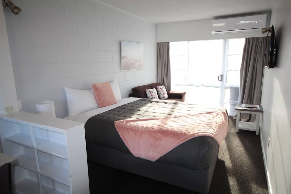 A hotel room with a bed, a desk, a chair, a window with curtains, and an air conditioning unit.