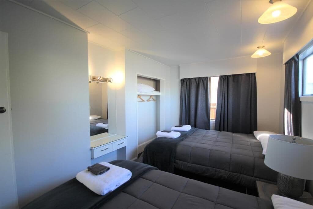 A hotel room with two beds, a bedside table with a lamp, a wardrobe, and a mirror.