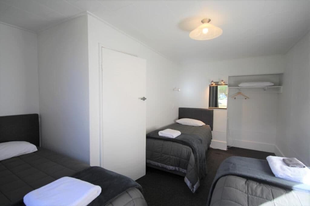 A hotel room with three single beds, a white door, a small window, and a shelf with a towel.