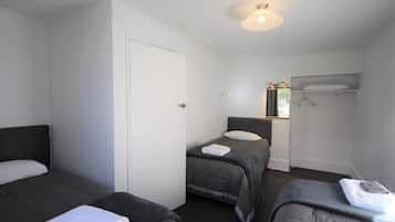 A hotel room with three single beds, a white door, a small window, and a shelf with a towel.