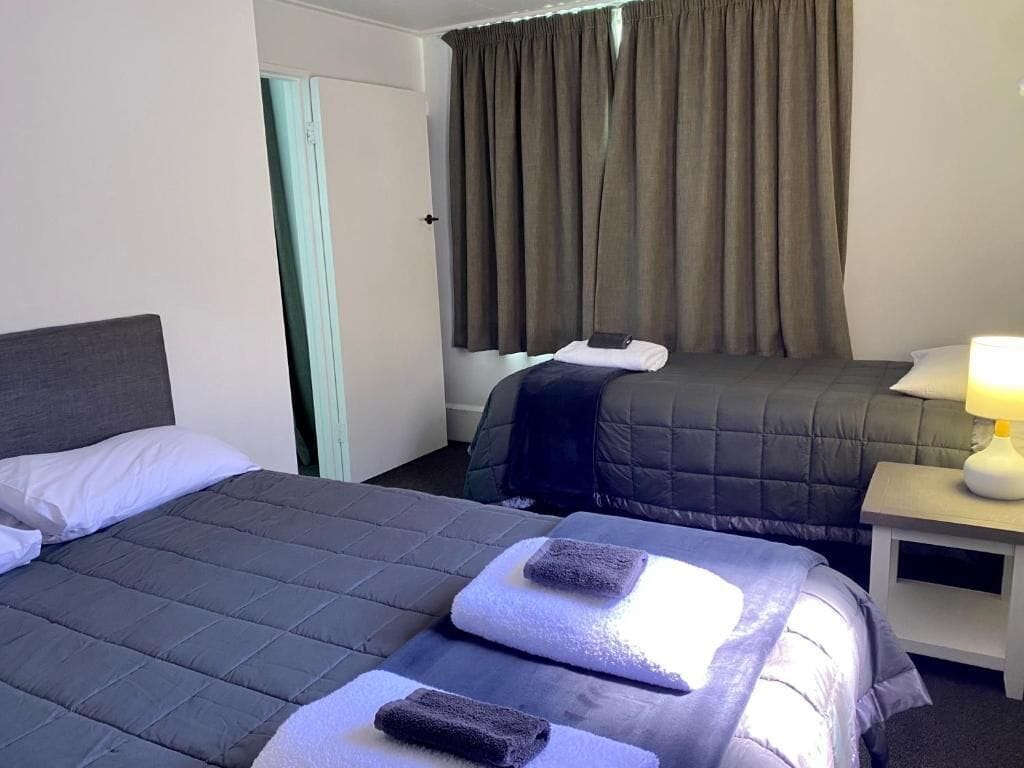 Una habitación de hotel con dos camas, una mesita de noche con lámpara y una ventana con cortinas.