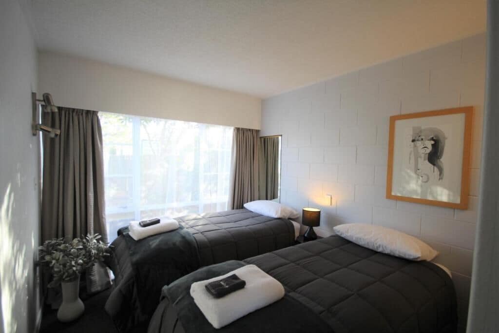 Una habitación de hotel con dos camas, una ventana con cortinas, un cuadro colgado en la pared y una mesita de noche con lámpara.