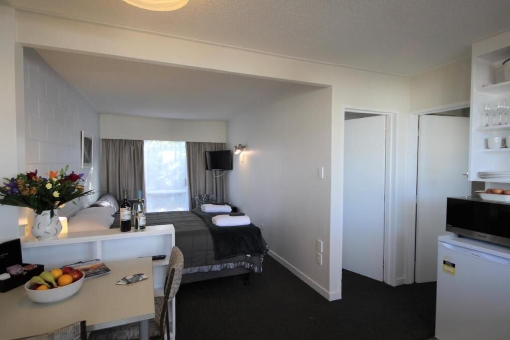 A compact hotel room with a bed, a desk with a vase of flowers, a television, and a minibar.
