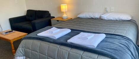 Habitación de hotel con una cama doble, una mesita de noche con lámpara, una silla y una pequeña mesa con un libro.