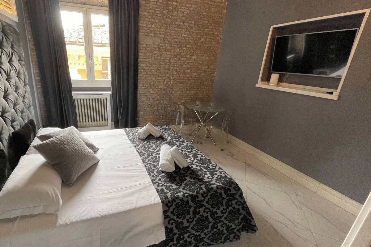 A hotel room with a tufted headboard, a bed with white linens, a patterned throw, and a small table with chairs.