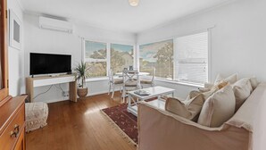 A modern living room with a flat-screen TV, a dining table with chairs, a sofa, and large windows with blinds.