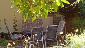 Een terras met metalen stoelen en een tafel, omgeven door begroeiing en een gevel.