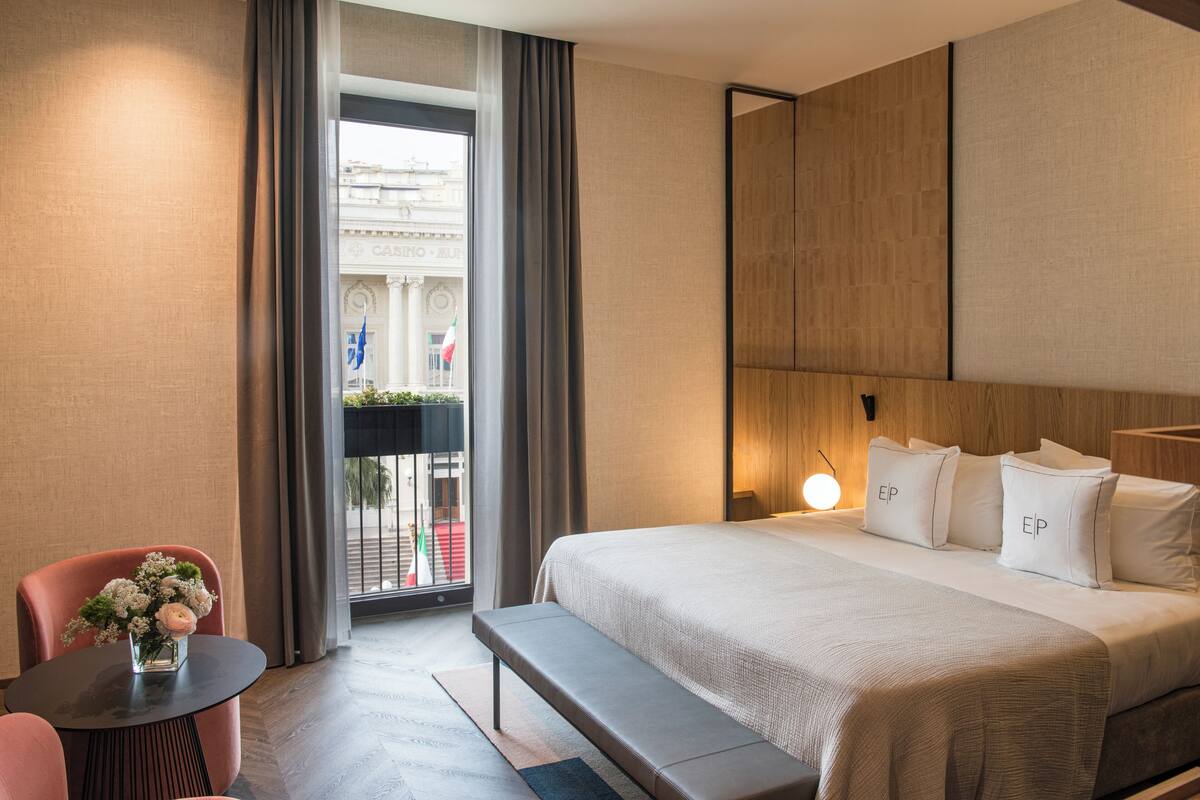 A hotel room with a large bed, a small table with a vase of flowers, a bench, and a view of a building with flags.
