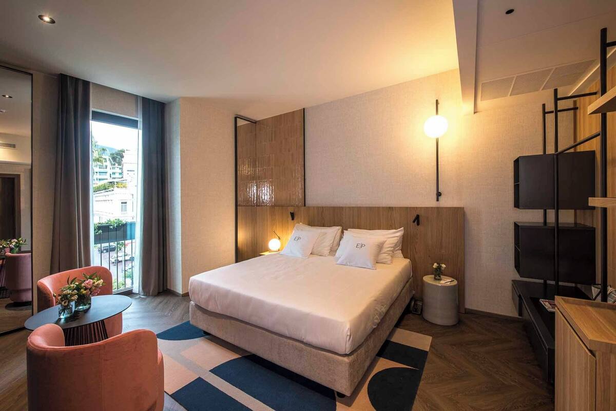 A modern hotel room with a large bed, a desk, a chair, and a view of the city.