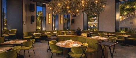 A restaurant with round tables, green upholstered chairs, and a large hanging light fixture.