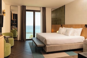 Habitación de hotel con una cama grande, un banco, un escritorio, una silla y vista al mar.