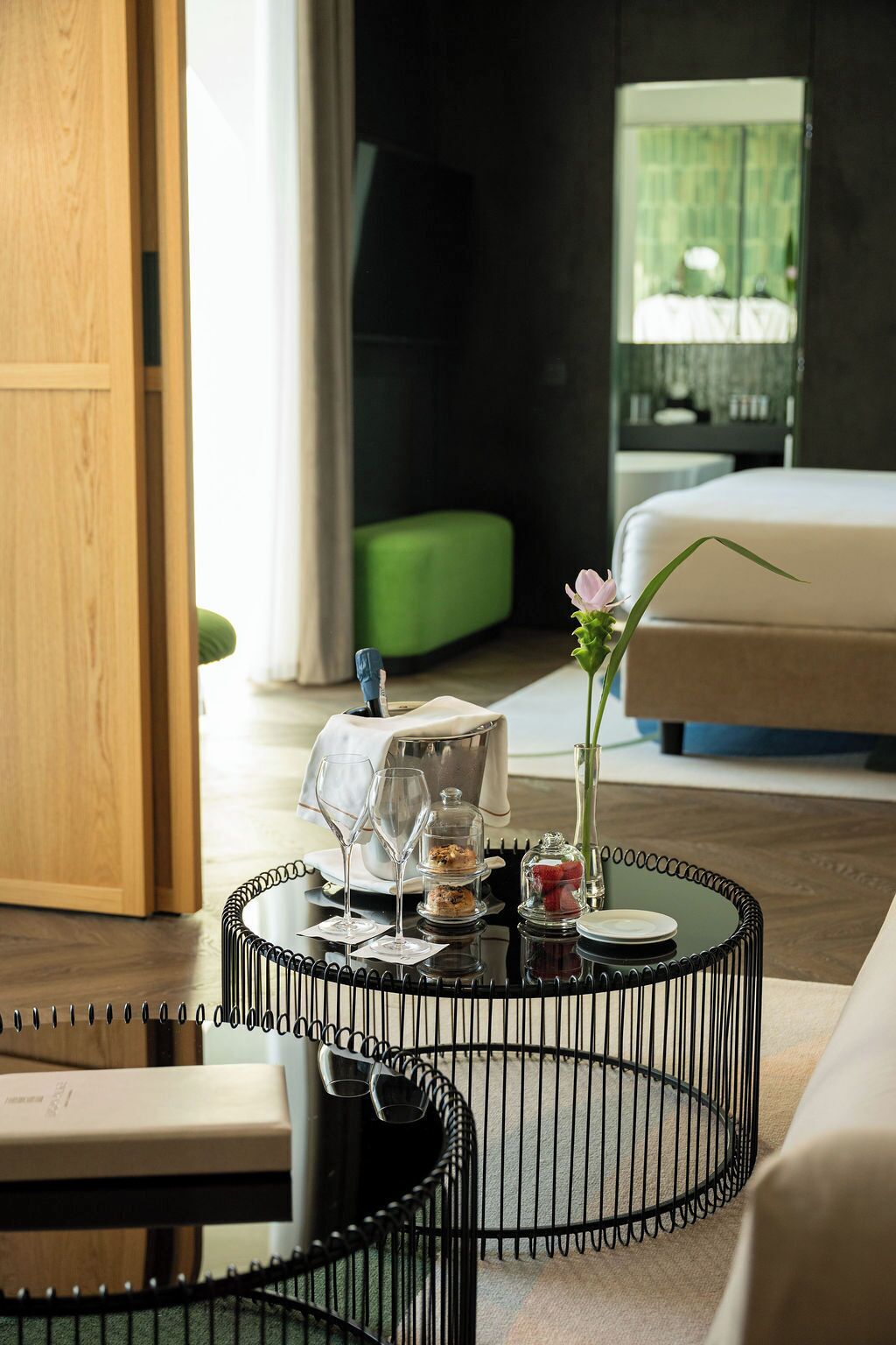 Une table à manger élégamment dressée, recouverte d’un cloche de verre, un vase contenant une seule fleur et une petite assiette, le tout installé dans une chambre d’hôtel moderne avec un pouf vert et un lit en arrière-plan.