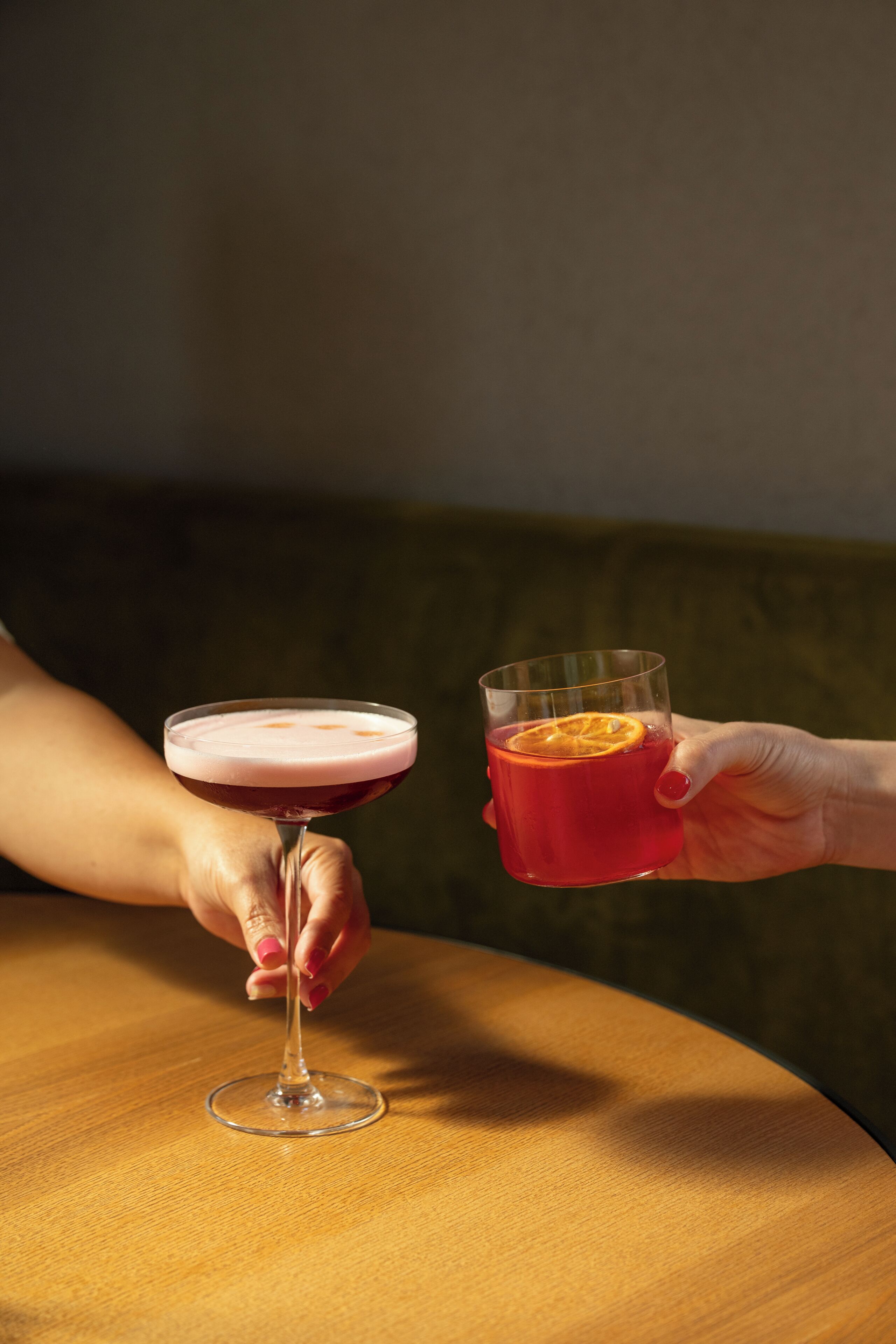 Two hands holding glasses of a beverage, one with a pinkish froth on top, the other with an orange slice.