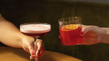 Two hands holding glasses of a beverage, one with a pinkish froth on top, the other with an orange slice.