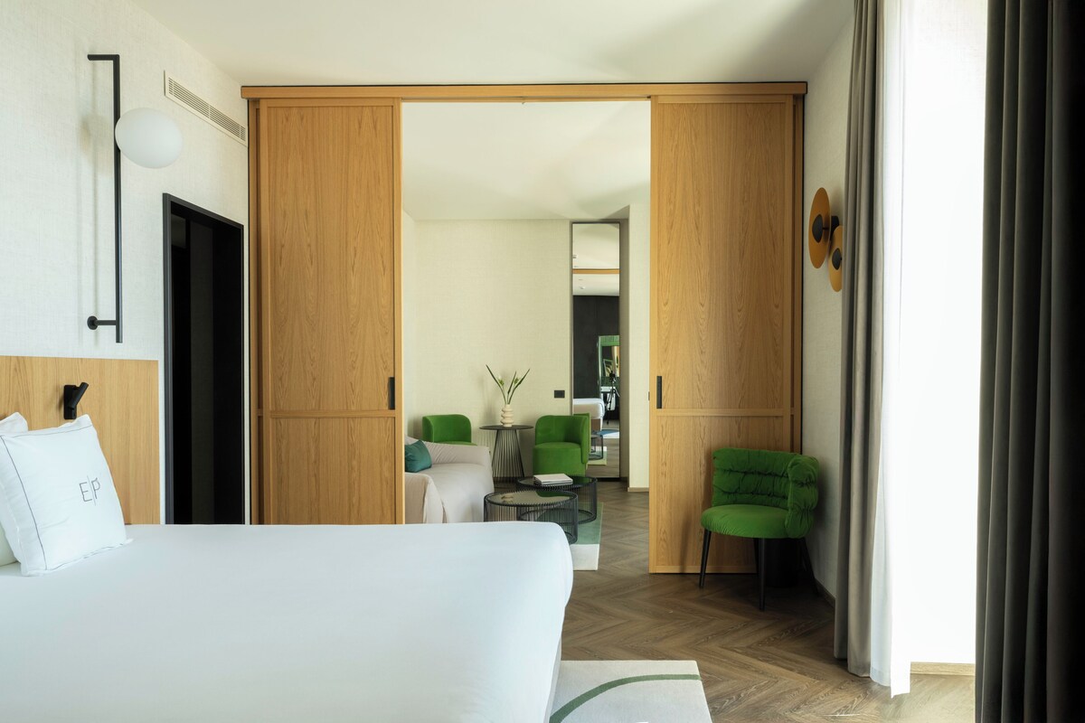 Une chambre d’hôtel comprenant un lit, un bureau, deux chaises, une petite table et une vue sur un espace repas accessible par une porte ouverte.