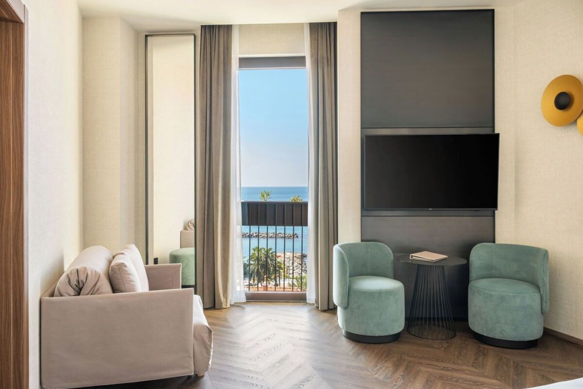 Un salon moderne avec un canapé, deux fauteuils, une petite table ronde et un téléviseur fixé au mur. Une grande fenêtre offre une vue sur la mer.