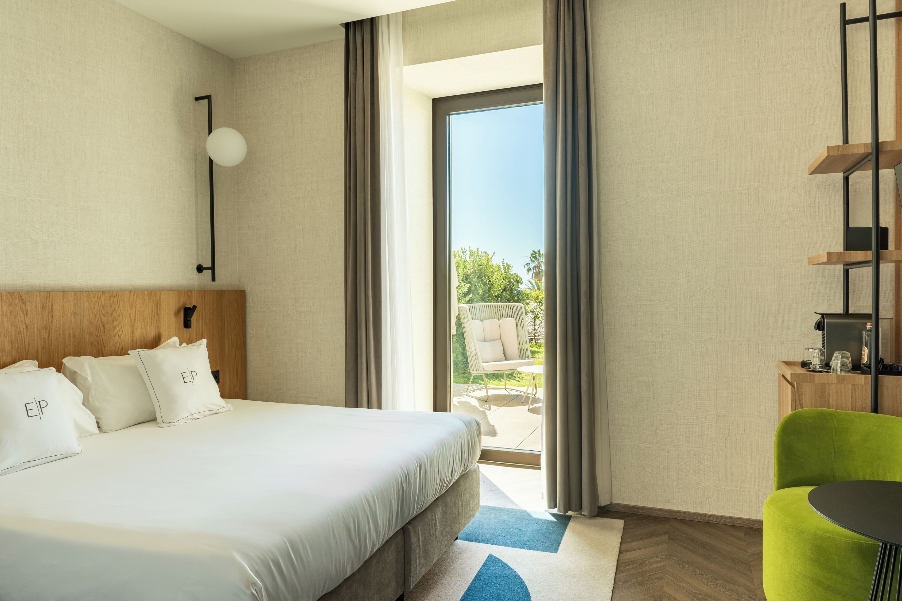 A hotel room with a large bed, a green chair, a small table, and a view of the outdoors.
