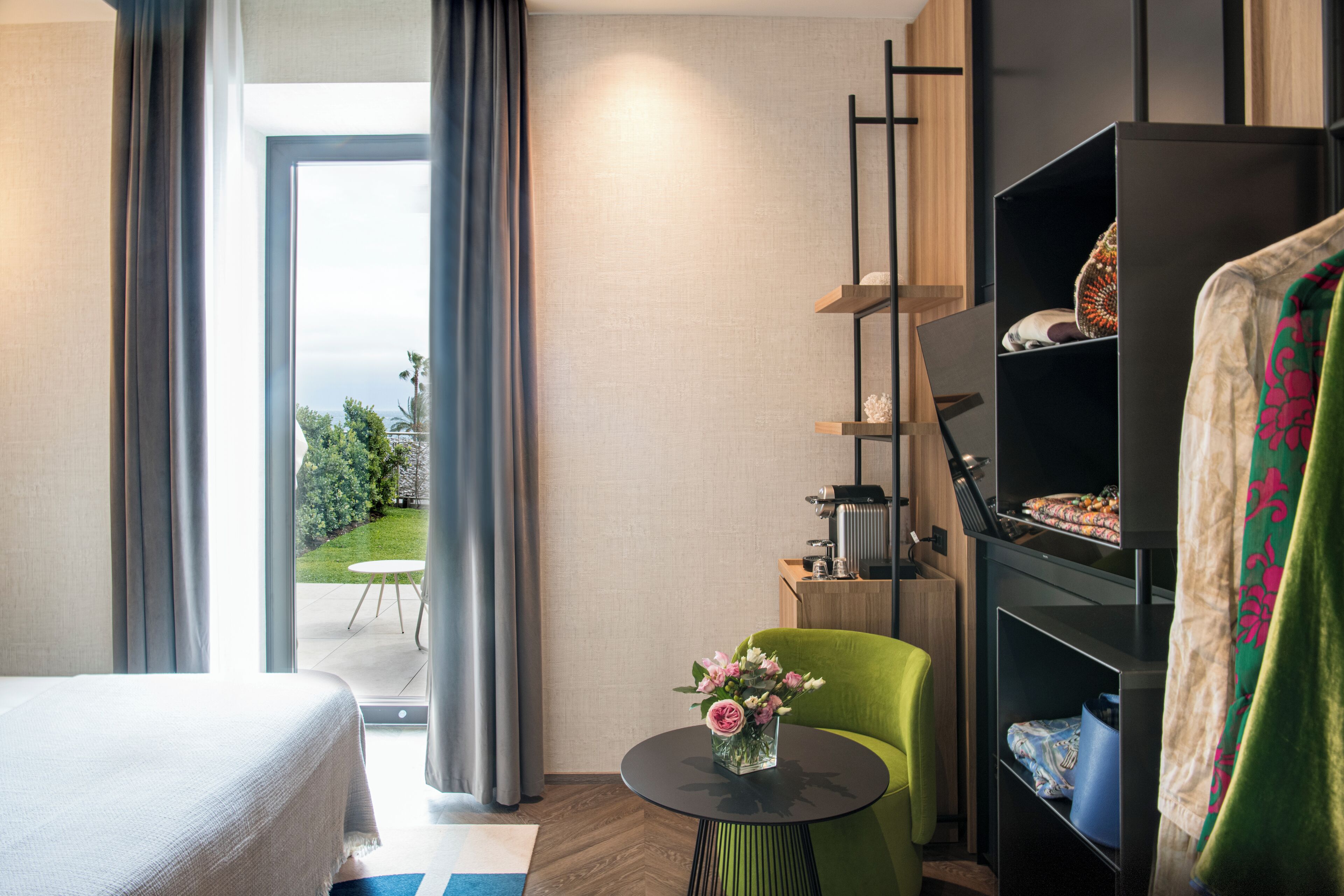 Camera d'albergo con un letto, un tavolino rotondo con un vaso di fiori, una poltrona verde, un mobiletto nero con vari oggetti e vista su un giardino attraverso un'ampia finestra.