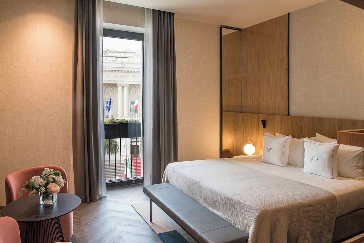 A hotel room with a bed, a bench, a small table with a vase of flowers, and a view of a building through a window.
