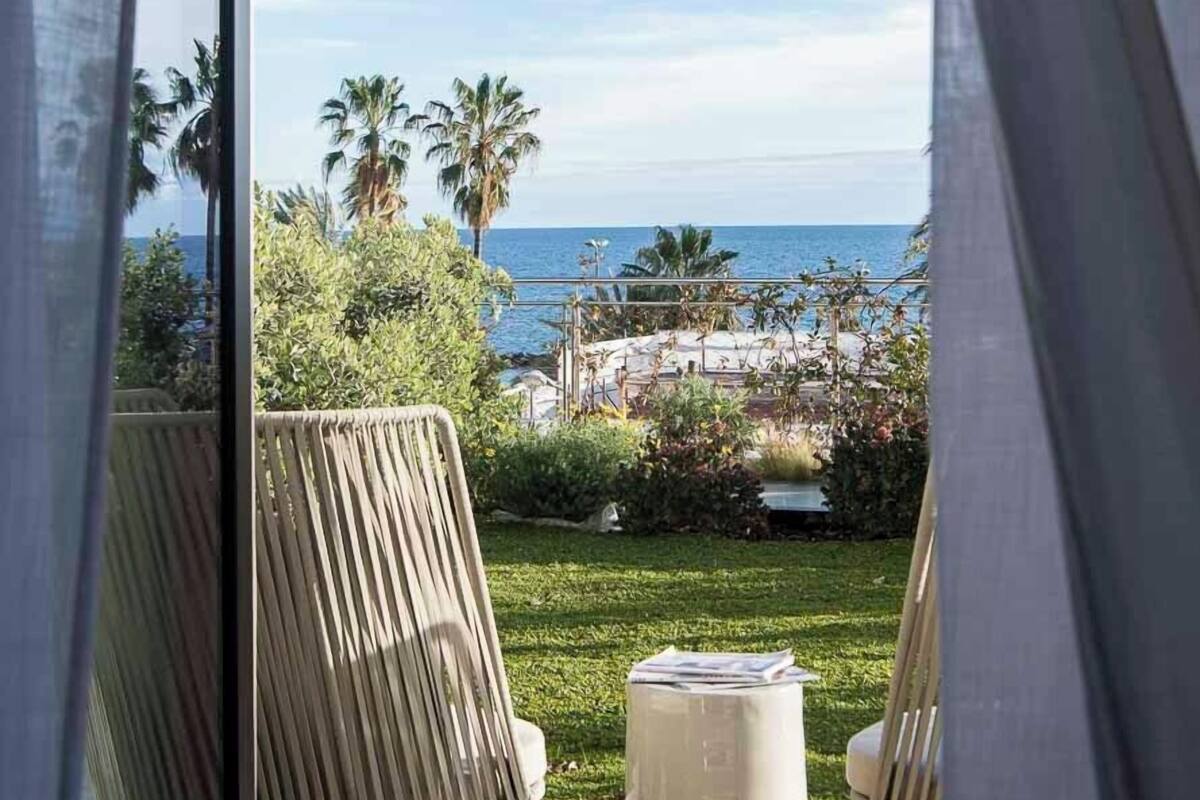 A balcony with two chairs and a small table, overlooking a beach with palm trees and a clear sky.