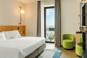 Camera d'albergo con un letto grande, vista tramite una porta vetrata, una poltrona verde e un mobile TV.