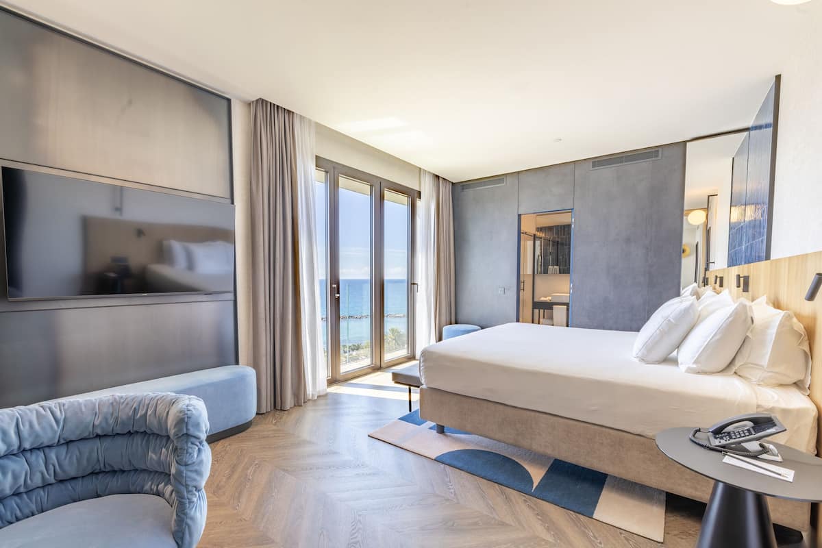 A modern hotel room with a bed, a sofa, a small table, and a large window offering a view of the sea.