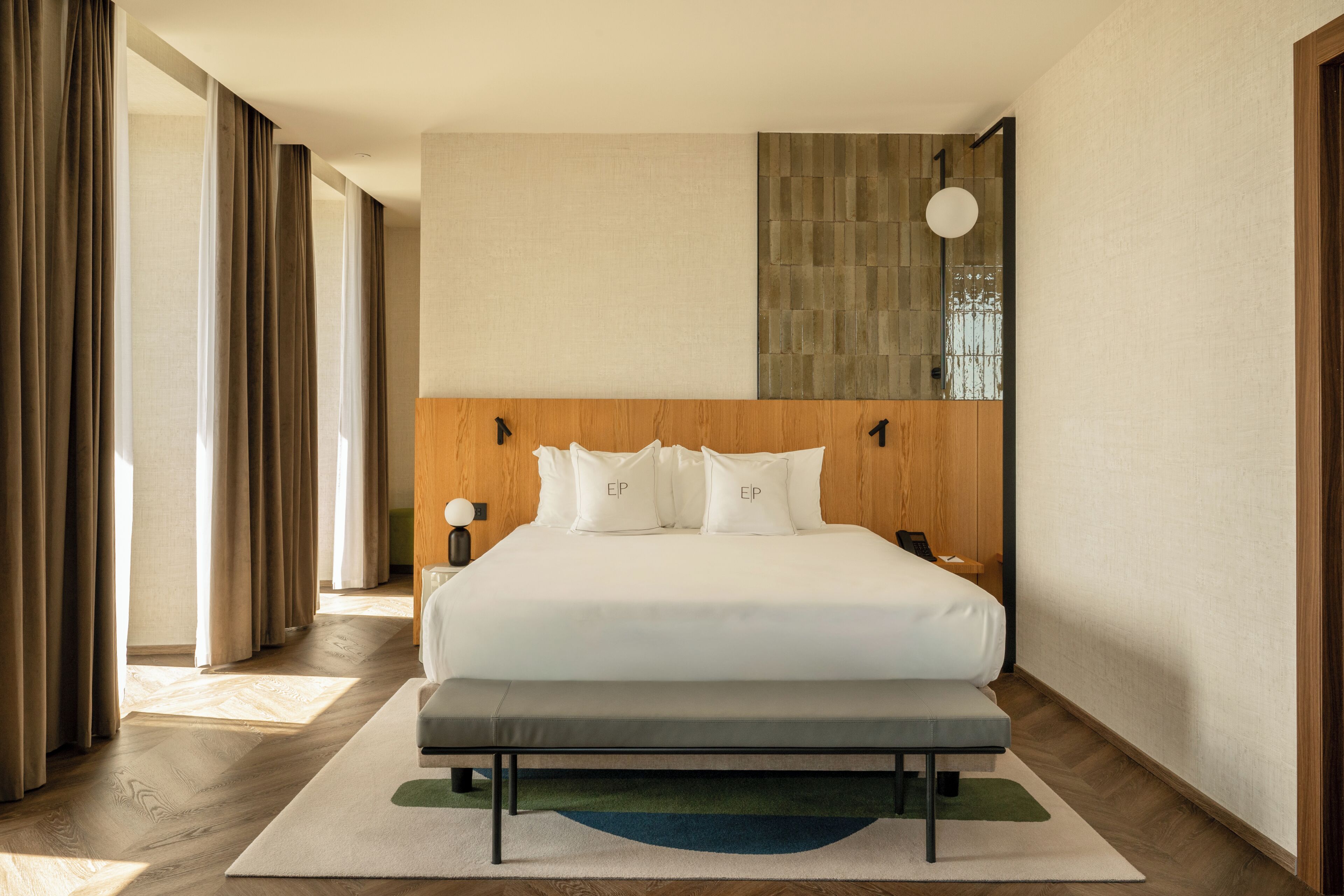 A hotel room with a large bed, a bench, a bedside table with a lamp, and a view of the outside through the window.