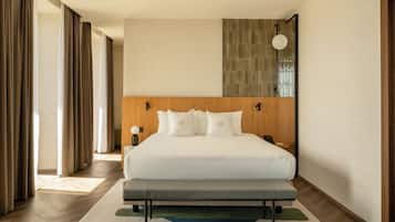 A hotel room with a large bed, a bench, a bedside table with a lamp, and a view of the outside through the window.