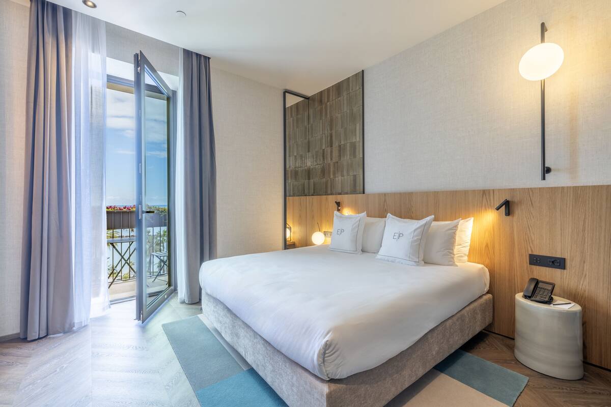 A modern hotel room with a large bed, a wooden headboard, a bedside table with a phone, and a view of the outdoors through a sliding door.