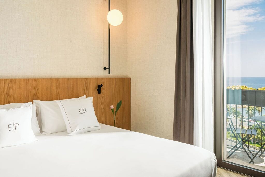 A hotel room with a bed, pillows with 'EIP' text, a nightstand, a wall-mounted light, and a view of the outdoors.