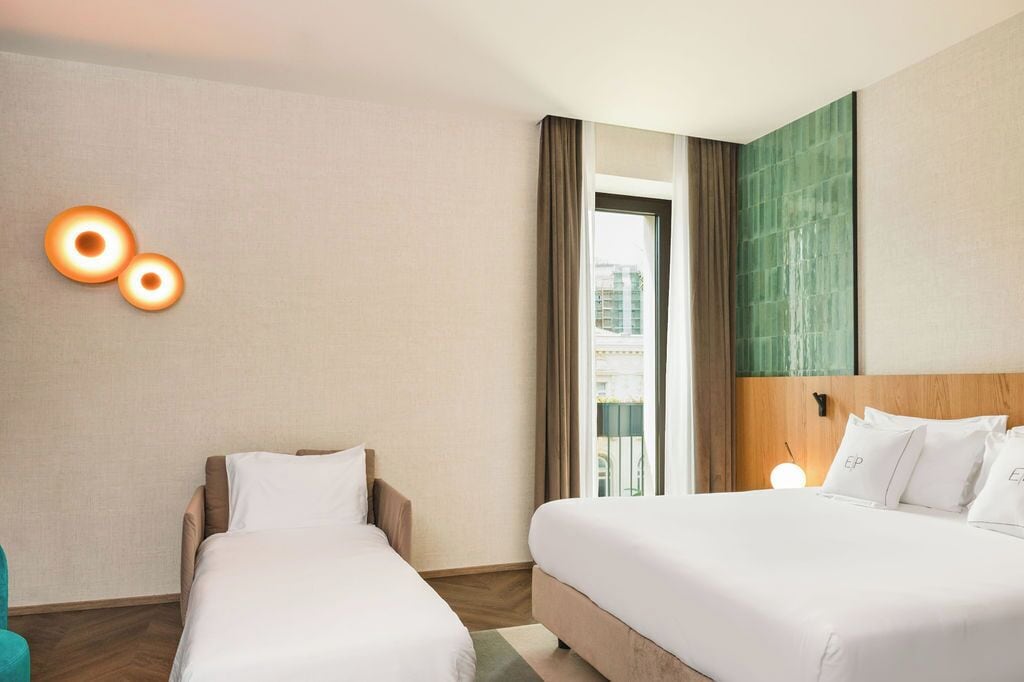Habitación de hotel con dos camas, una mesita de noche con lámpara, vista a la ciudad por una ventana y una lámpara de pared.