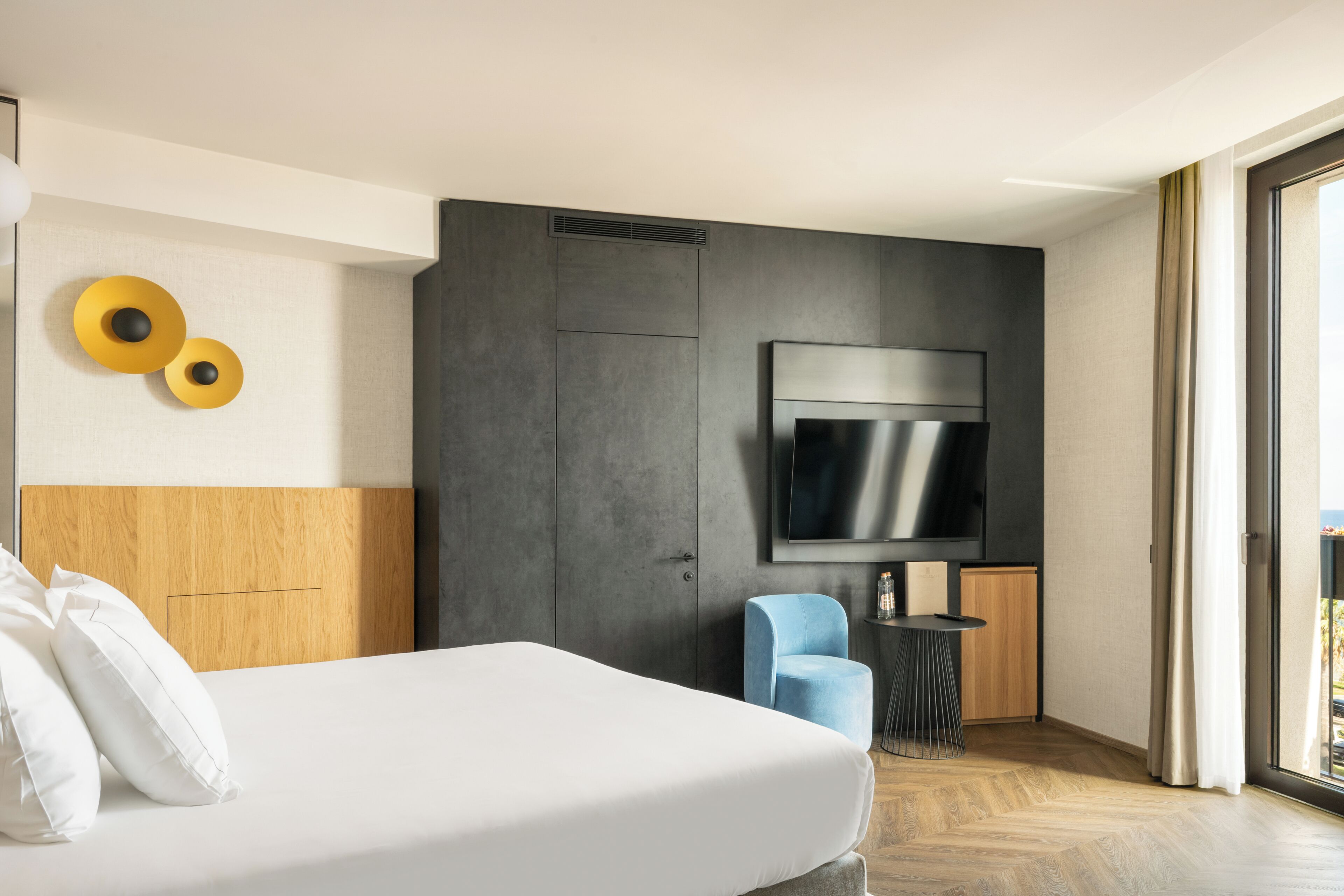 A modern hotel room with a large bed, a flat-screen TV mounted on a dark wall, a small round table with a chair, and a view of the outdoors through a large window.