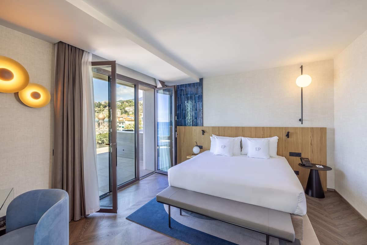 A modern hotel room with a large bed, a desk, a chair, and a view through the glass door.