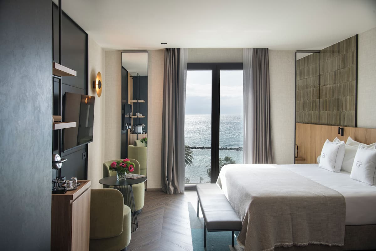 A modern hotel room with a large bed, a desk, a chair, and a view of the sea.
