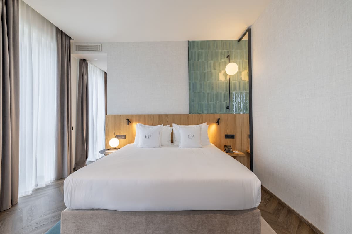 A hotel room with a large bed, a nightstand with a lamp, a wall-mounted light, and a view of the outdoors through the window.
