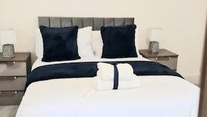 Una cama bien hecha con edredón blanco y almohadones azul marino, un cabecero gris y dos mesitas de noche con lámparas.