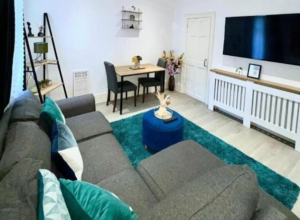 A living room with a sectional sofa, a dining table with chairs, a blue ottoman, and a flat-screen TV mounted on the wall. - Strood Home-sleeps 6-great for Groups & Families (Rochester)