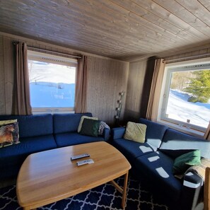 A cabin interior with a blue sofa, a wooden table, and large windows with a snowy view. - Captivating 6-bed Cottage in Lismarka (Ringsaker)