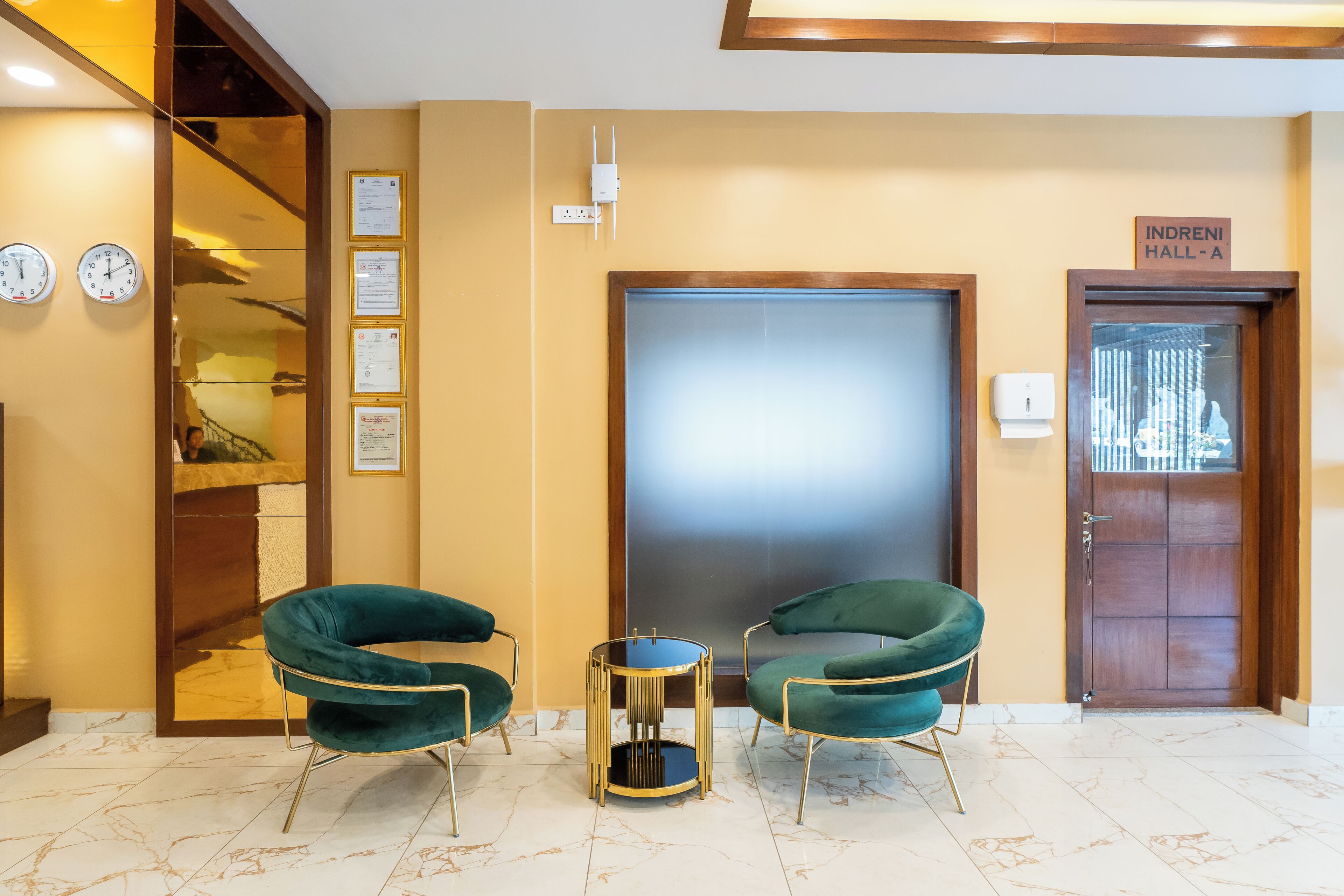 A reception area with two green armchairs, a small round table, a wooden door labeled "INDRENI HALL-A", and a wall with clocks and framed certificates.