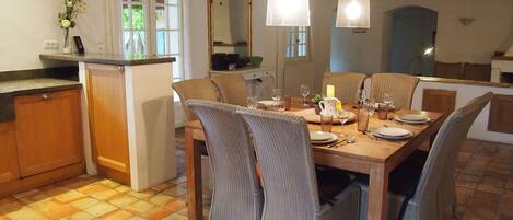 A dining area with a wooden table set for four, surrounded by wicker chairs. There is a small kitchen area with a refrigerator and a counter.